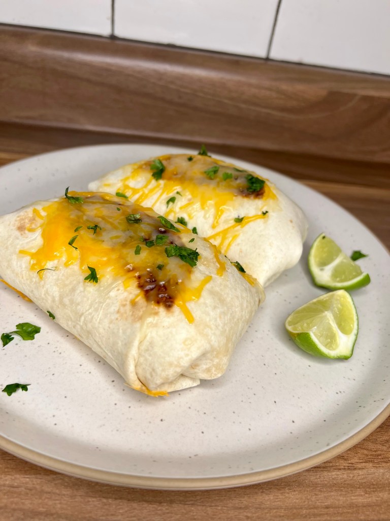 Two burritos with melted cheese on the top on a white plate with some cilantro and lime wedges