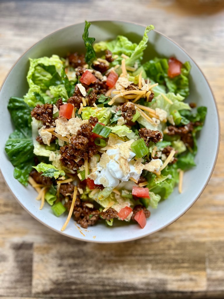 Lettuce, tomato, onion, cilantro, cheese, taco meat, sour cream, and tortilla chips in a white bowl