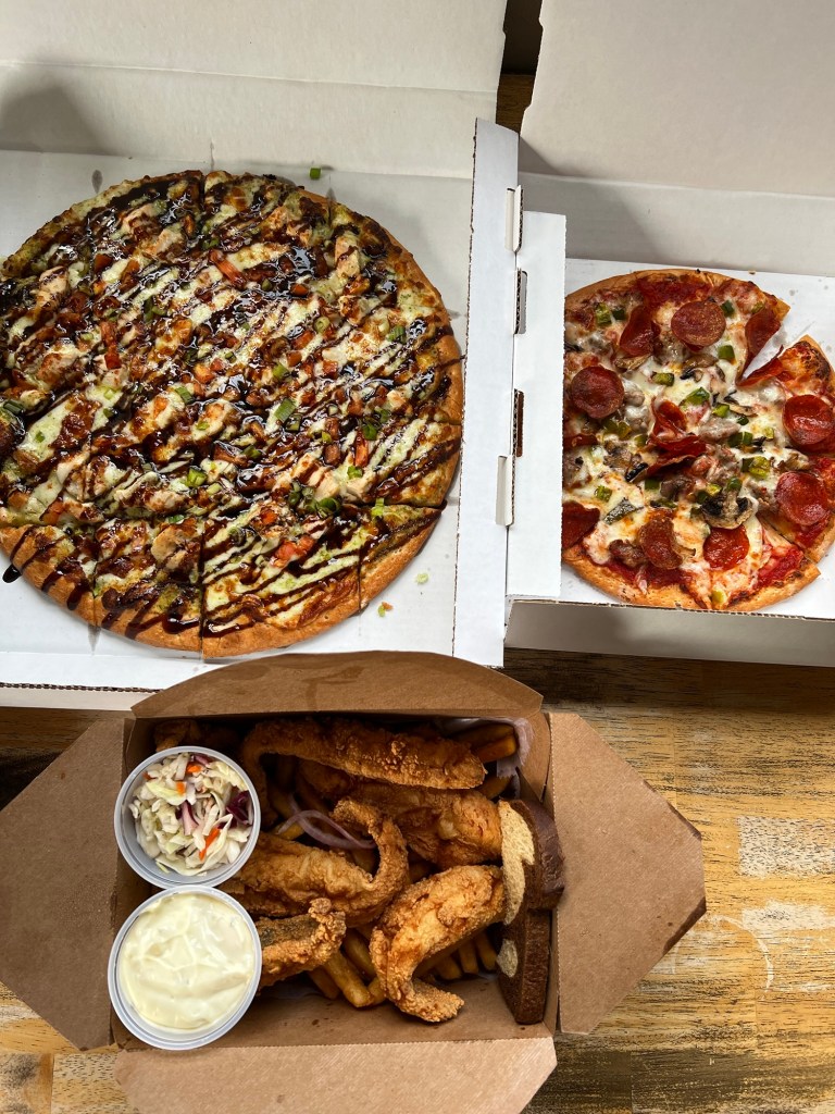 Two pizzas in a box with a box of fish and fries below on a wood table