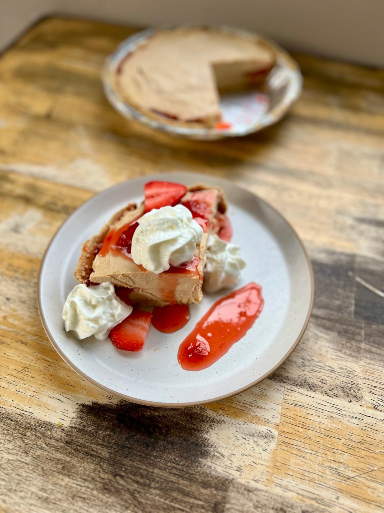 A Peanut Butter and Jelly Pie on a white plate with whipped cream, compote, and strawberry on top on a wooden table