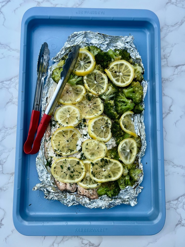 Salmon and broccoli foil pack opened up on a blue baking sheet after just being grilled