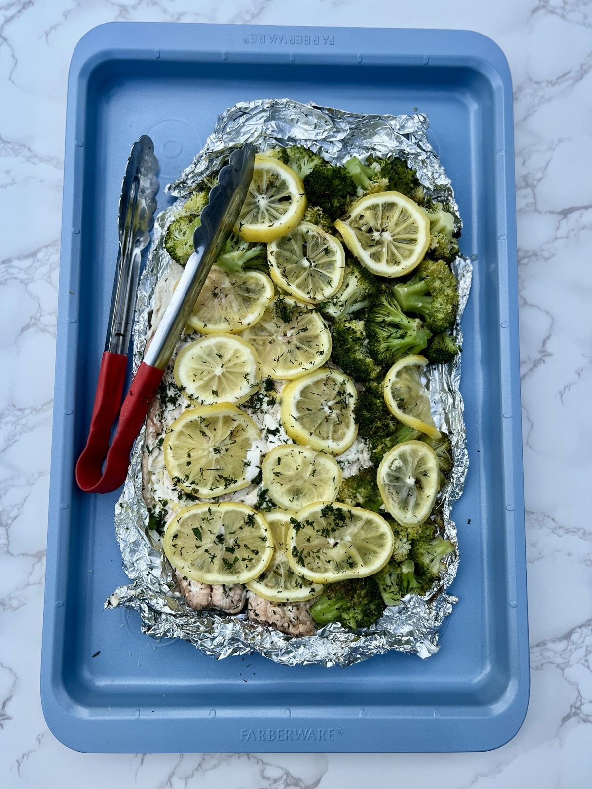 A foil pack with salmon and broccoli on a blue baking sheet with a tong sitting on it