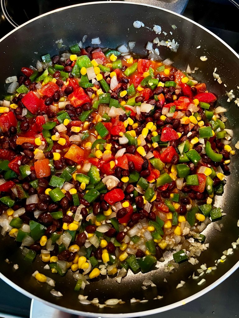 A skillet filled with pepper, onion, garlic, tomato, corn, beans, and seasonings on the stove 