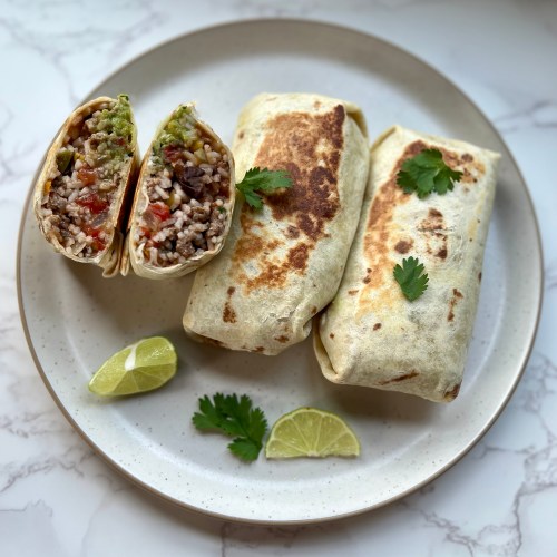 Three southwest burritos with one cut in half on a white plate with lime and cilantro on the side