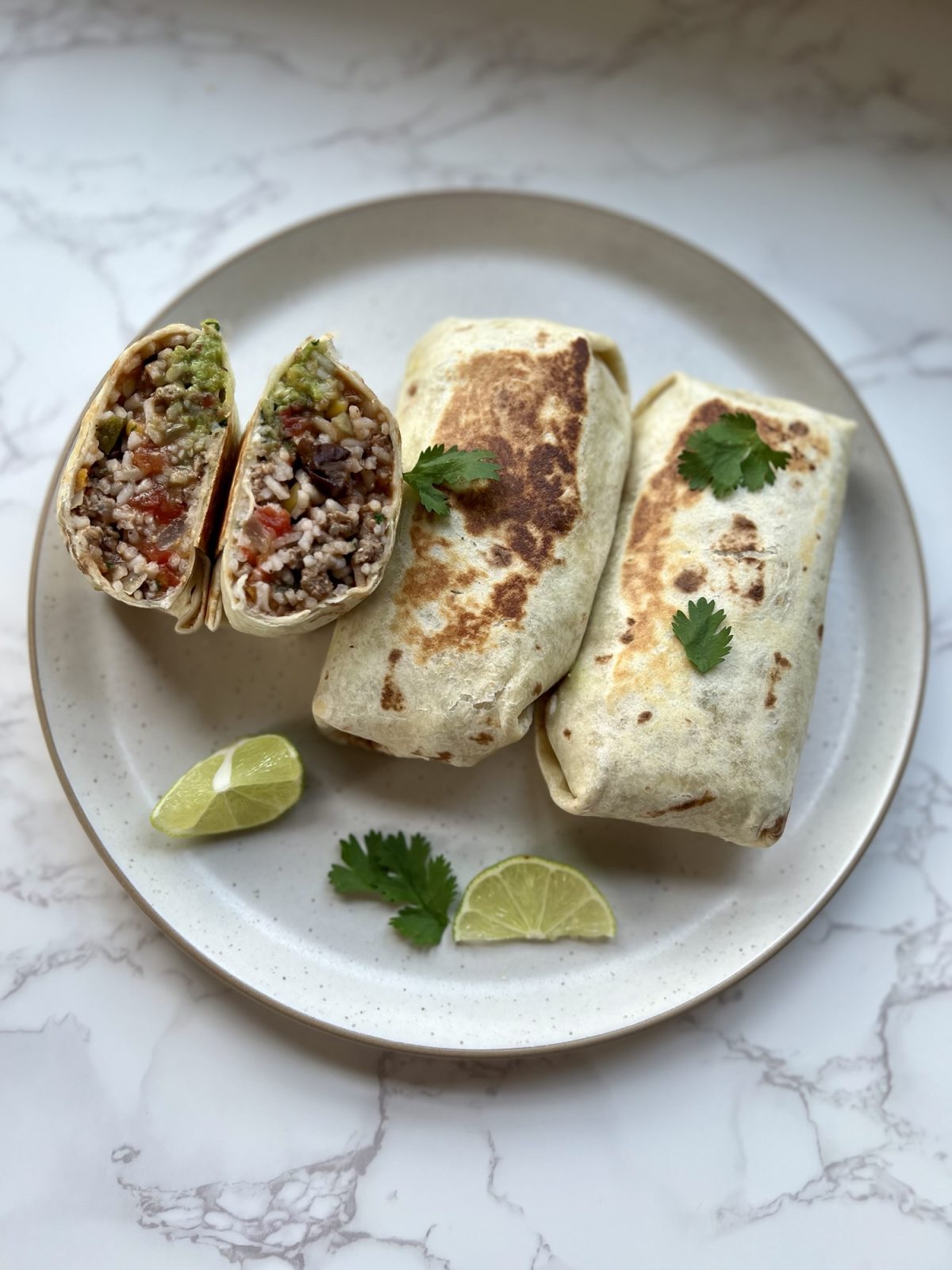 Three southwest burritos with one cut in half on a white plate with lime and cilantro on the side