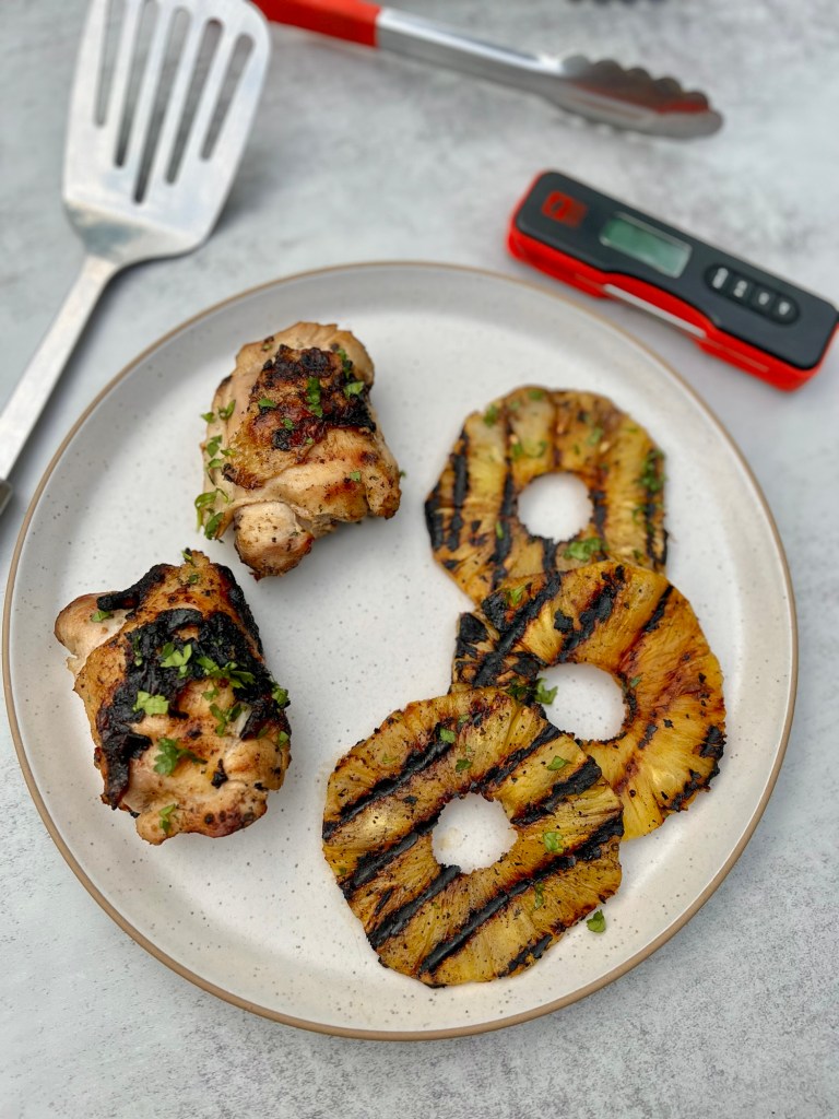 Hawaiian chicken thighs and grilled pineapple on a white plate with a spatula, tongs, and meat thermometer nearby