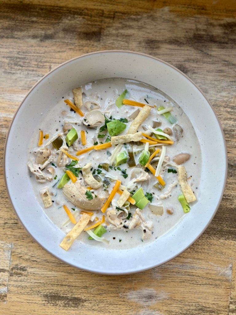 White Chicken Chili in a white bowl with cheese, cilantro, wonton strips, and avocado garnished on top