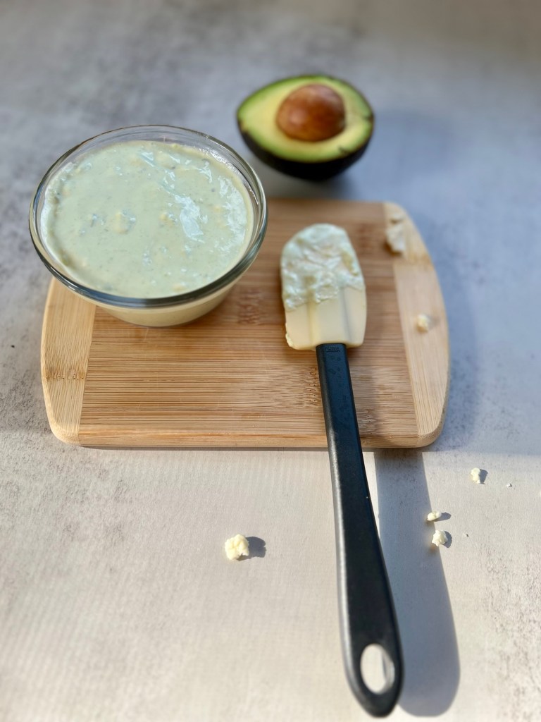 Avocado blue cheese dressing in a dish on a cutting board with a rubber spatula and avocado half nearby
