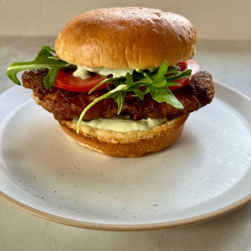A spicy fried chicken sandwich with tomato, arugula, and avocado blue cheese dressing on a white plate