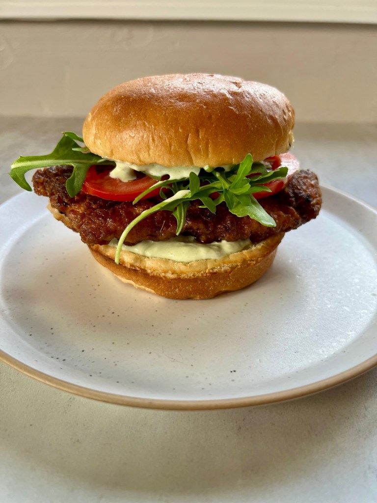 A spicy fried chicken sandwich with tomato, arugula, and avocado blue cheese dressing on a white plate