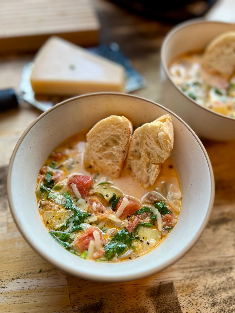 A bowl of soup with some baguette pieces inside with some more of it behind on a wooden table