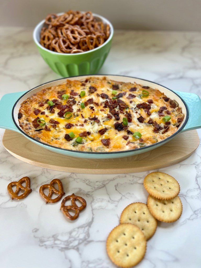 Bacon Jalapeño Popper dip in a glass dish with a bowl of pretzels behind it and some crackers laying around