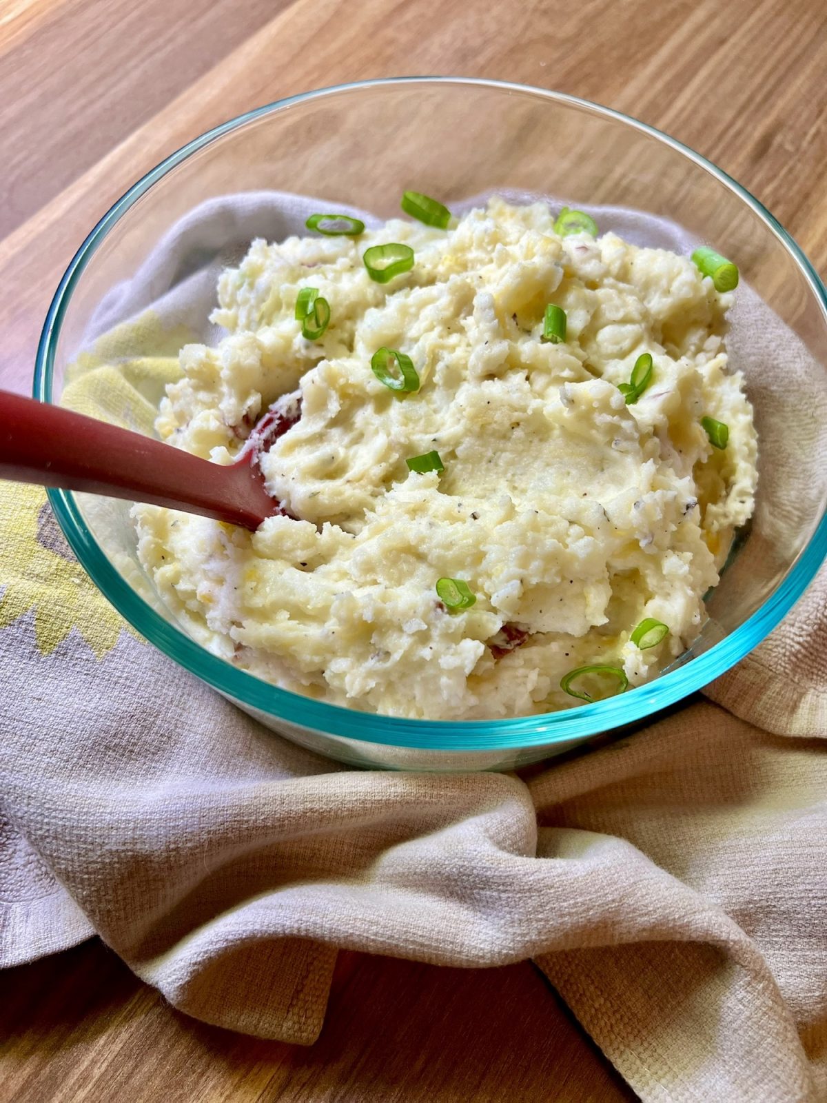 Creamy mashed potatoes in a glass dish with green onion on top and a red serving spoon inside