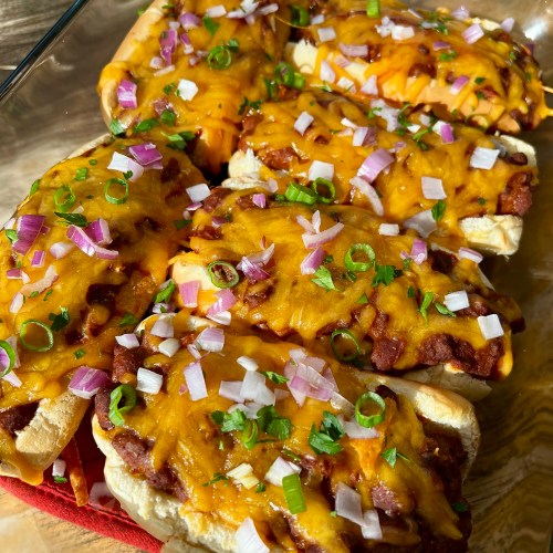 Chili cheese dogs in a glass baking dish with garnishes on top on a wooden table