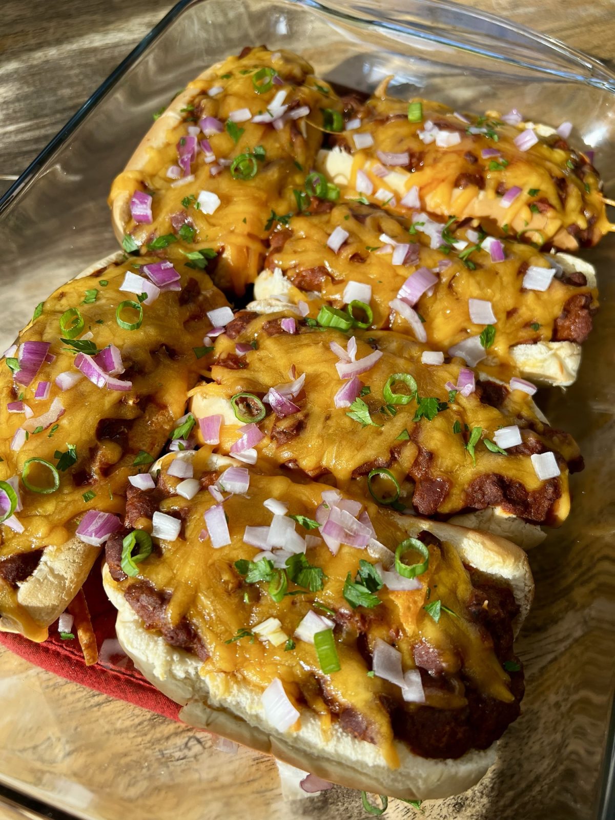 Chili cheese dogs in a glass baking dish with garnishes on top on a wooden table