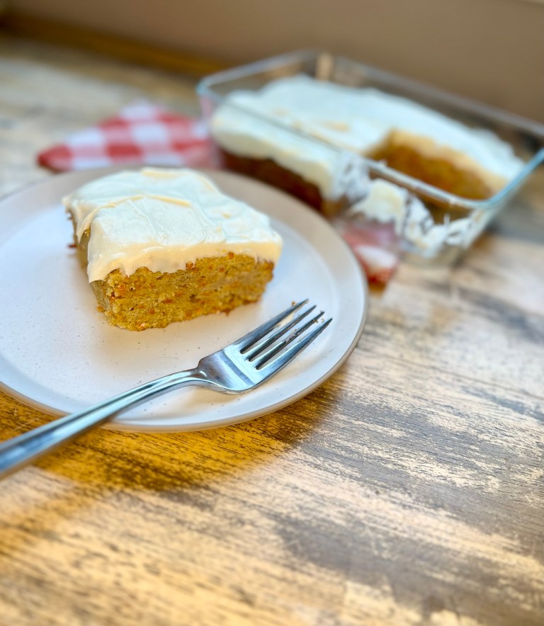 Carrot cake bars in a glass pan with a white plate in front of it with a carrot cake bar on it