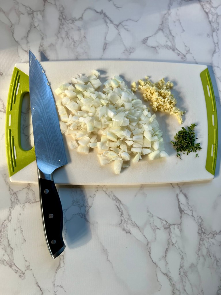 Onion, garlic, and thyme cut up on a cutting board with a knife nearby