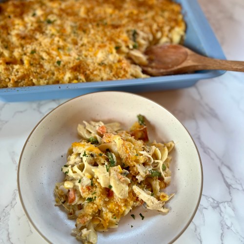Chicken noodle casserole in a white bowl with a blue pan of it behind with a wooden spoon inside