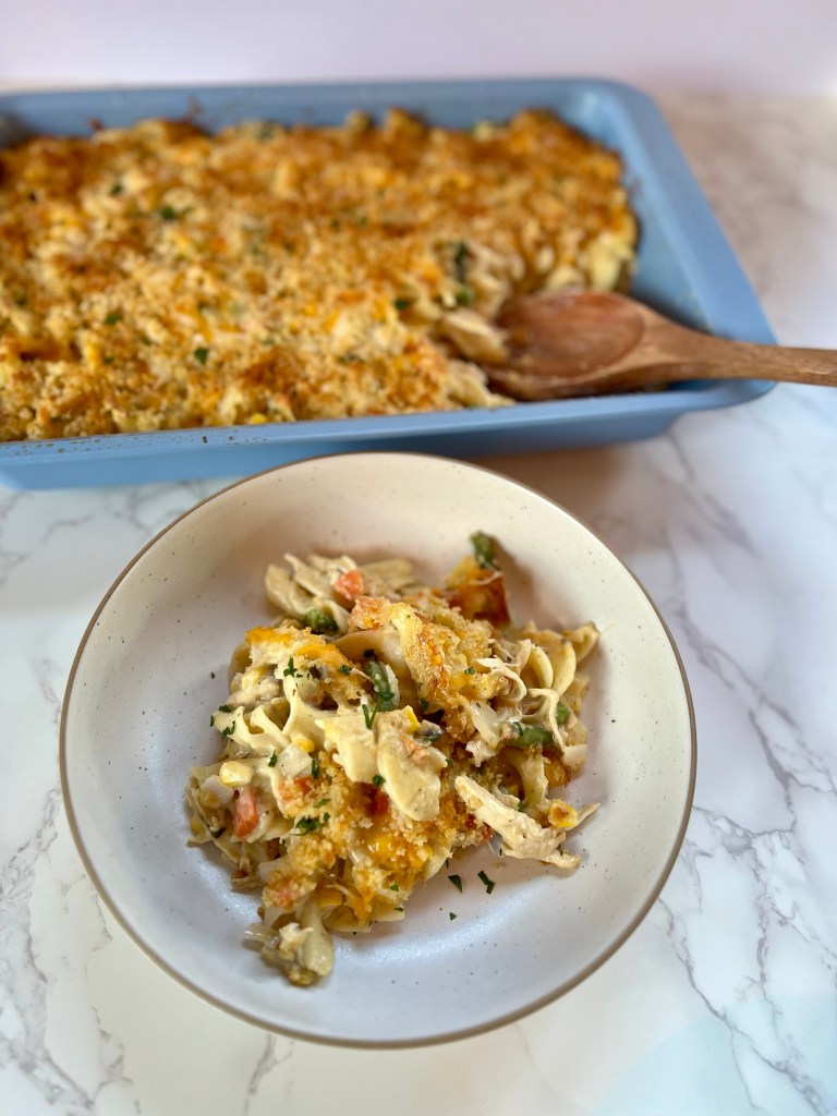 Chicken noodle casserole in a white bowl with a blue pan of it behind with a wooden spoon inside