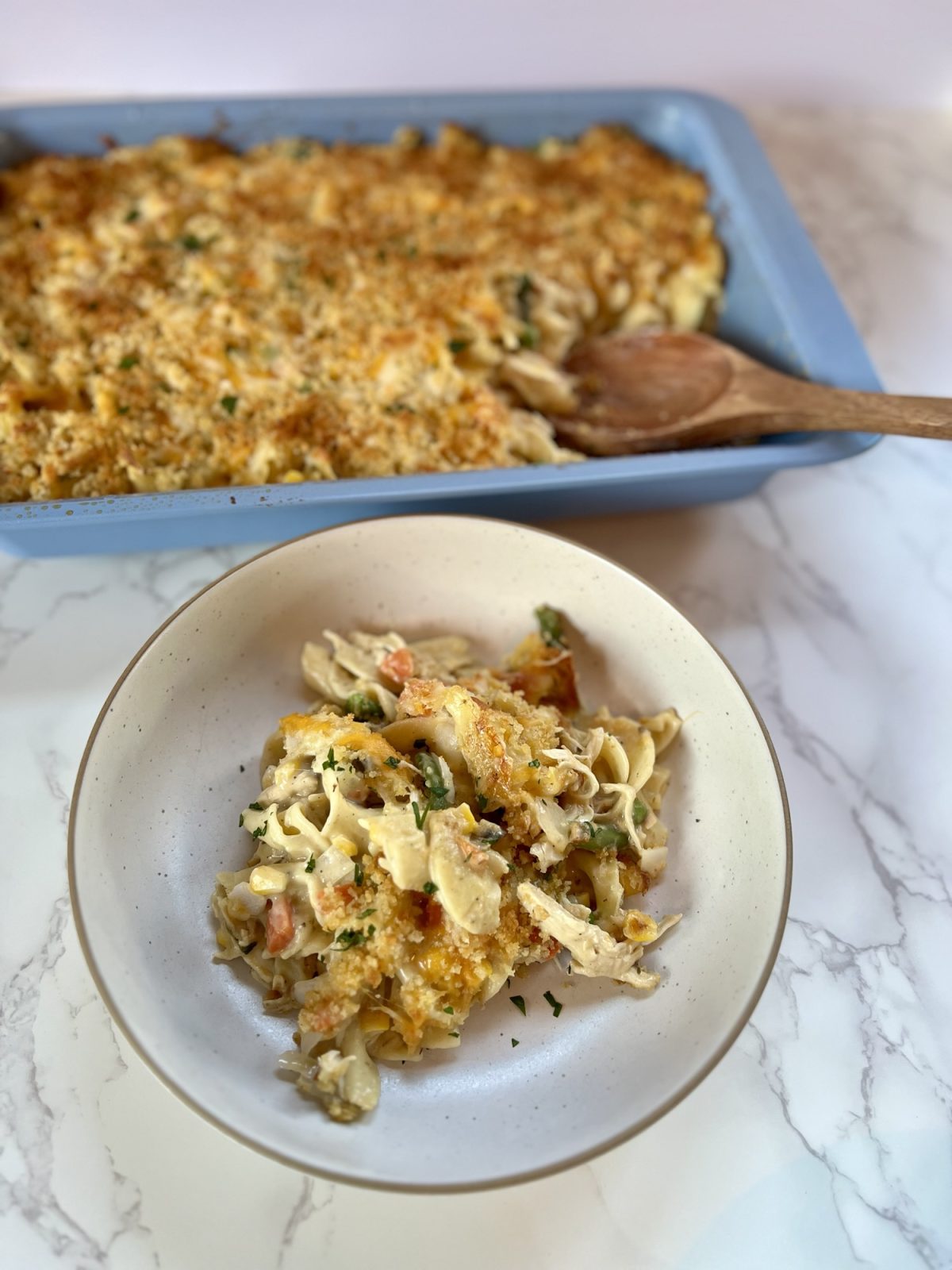 Chicken noodle casserole in a white bowl with a blue pan of it behind with a wooden spoon inside
