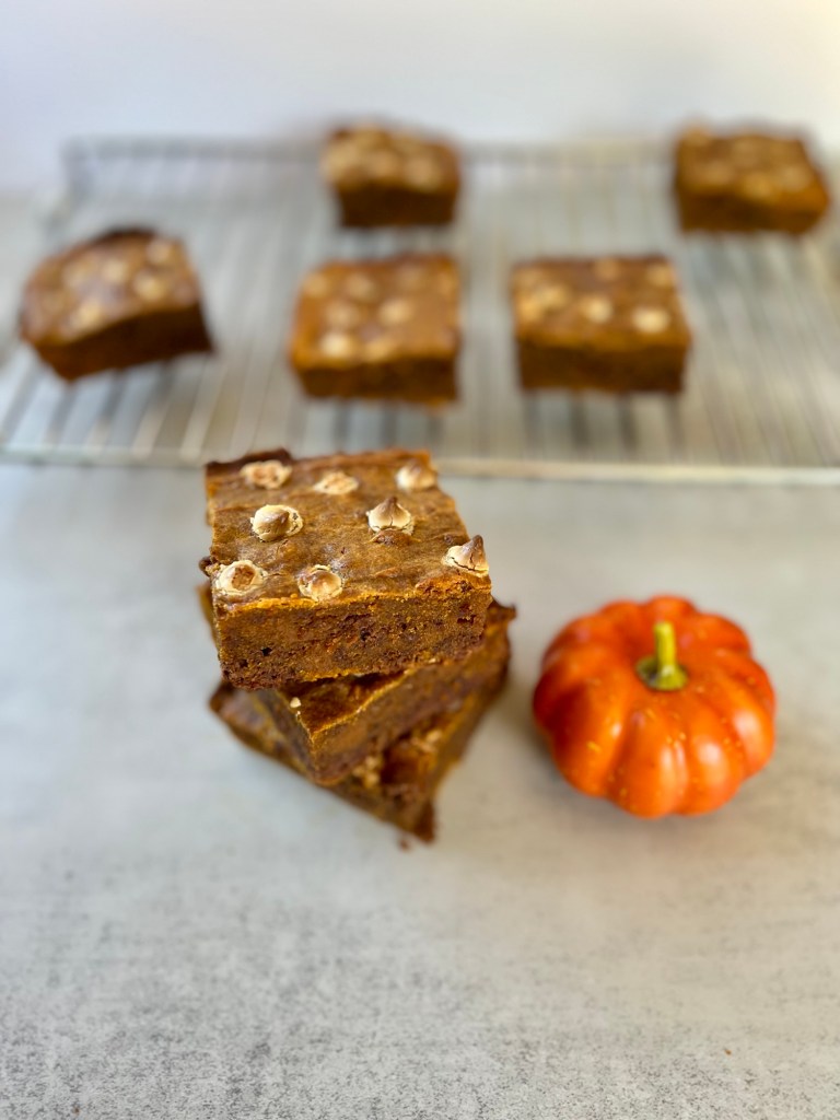 Three brownies stacked up with a pumpkin decoration next to it in front of a cooling rack with more brownies on it