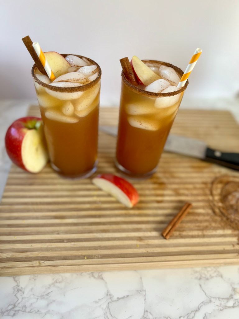 Two apple cider whiskey sours on a wooden cutting board with some apples, cinnamon sticks, and spices laying around