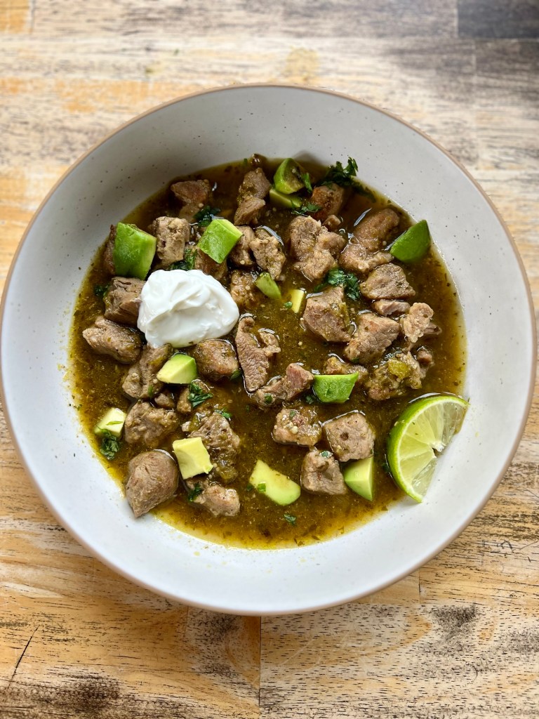 Chili Verde in a white bowl with sour cream, cilantro, avocado, and lime for garnish