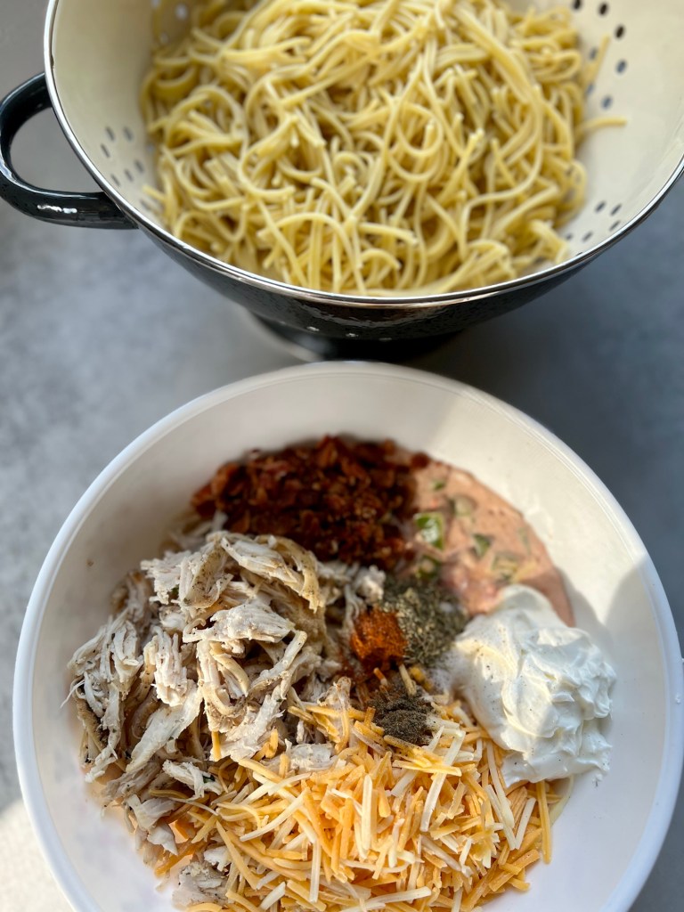 A large bowl filled with the filling for chicken spaghetti with a strainer filled with noodles behind it