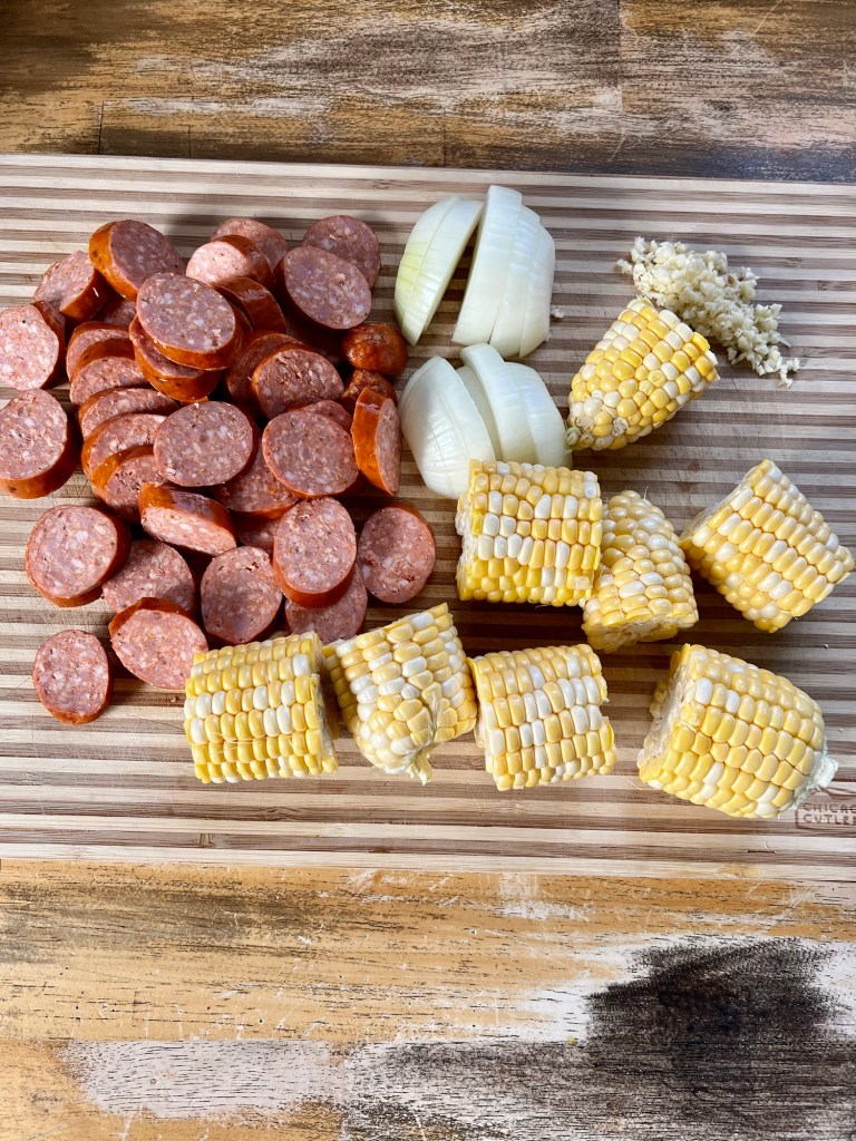 A cutting board with corn, onion, sausage, and garlic cut up on it