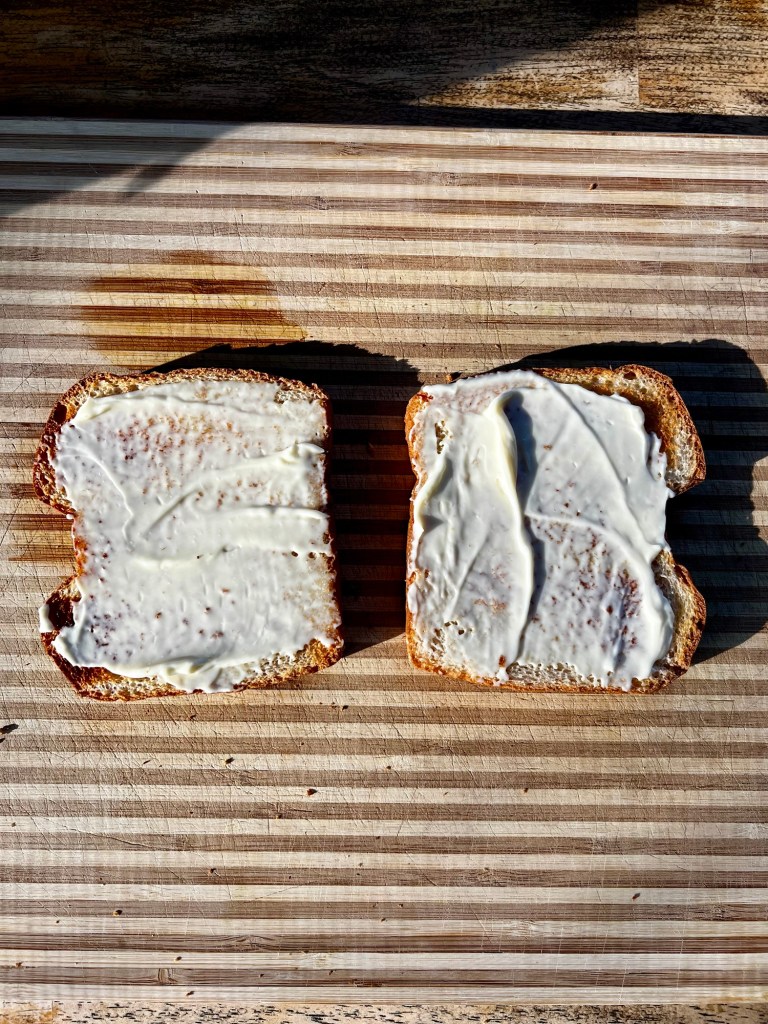 Two pieces of toast with mayo on them on a wooden cutting board