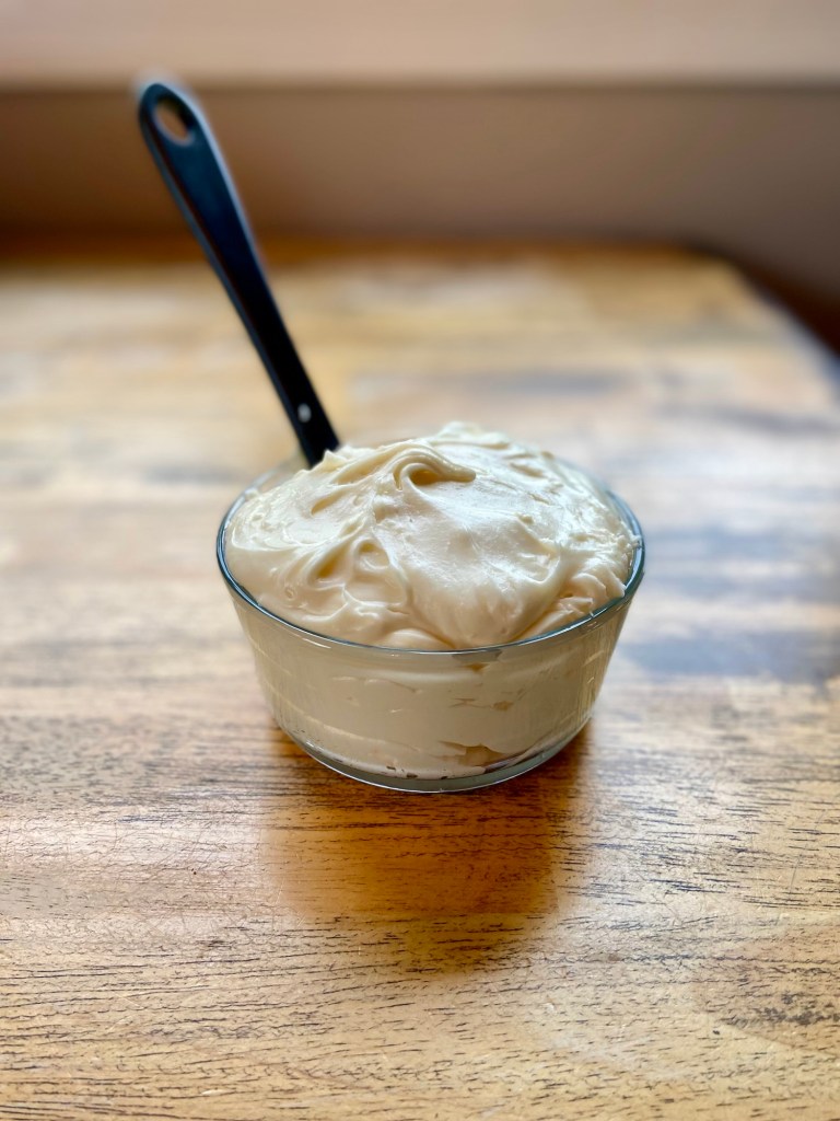 Cream cheese frosting in a glass dish with a rubber spatula sticking out of it, on a wooden table