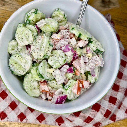 Creamy cucumber salad in a bowl with a red and white towel underneath it on a wooden table