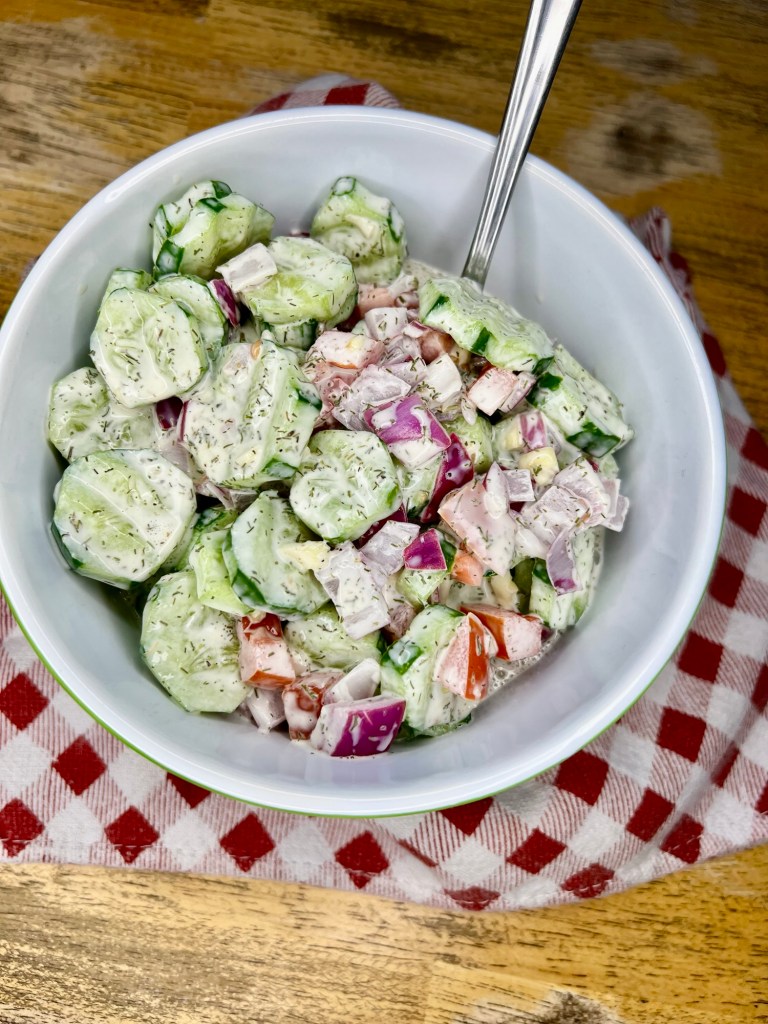 Creamy cucumber salad in a bowl with a red and white towel underneath it on a wooden table
