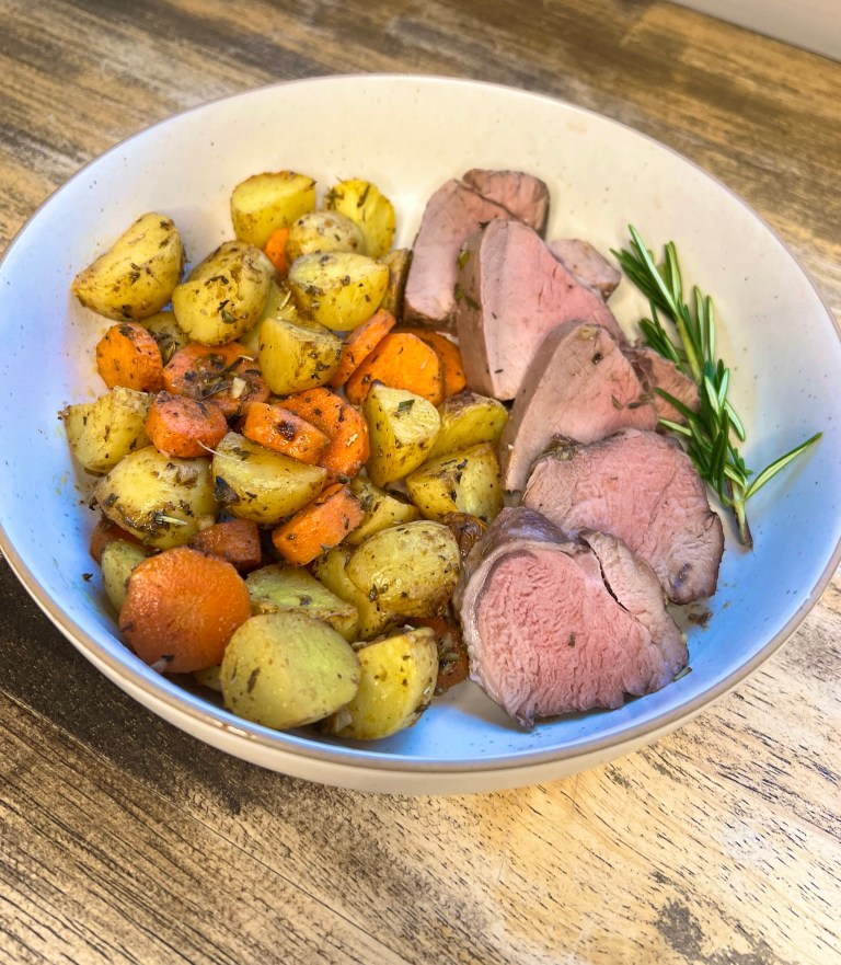 Pork tenderloin and veggies in a white bowl with some rosemary on the side, on a wooden table