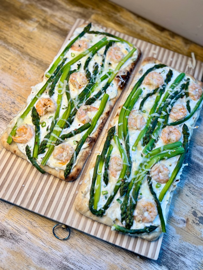 Two flatbreads with shrimp and asparagus on them on a wooden cutting board