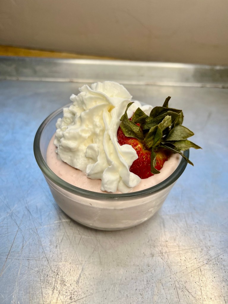 Strawberry mousse in a small dish with whipped cream and a strawberry on top, on a baking sheet