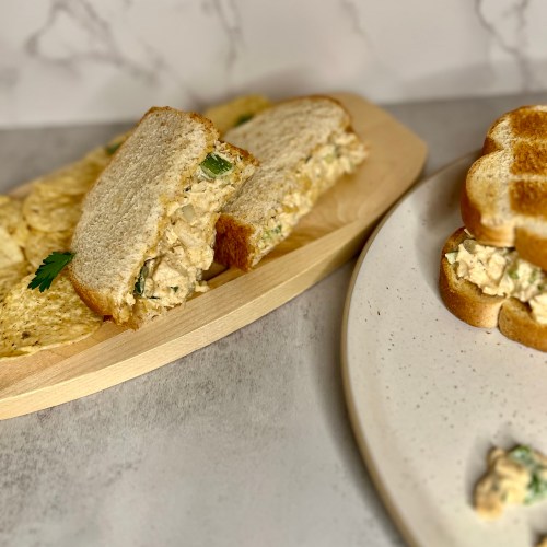 A chicken salad sandwich with some chips next to it on a wood board with a plate next to it