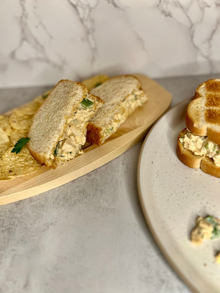 A chicken salad sandwich with some chips next to it on a wood board with a plate next to it