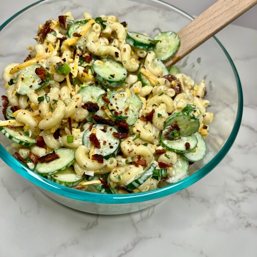 Cucumber Bacon Pasta Salad in a glass dish with a wooden spoon on a marble countertop