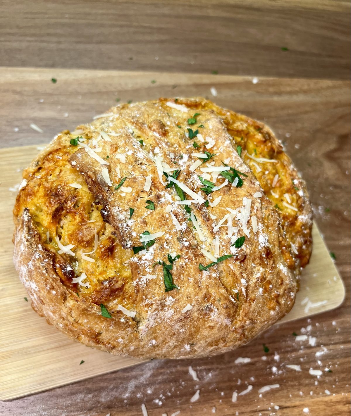 Herb and Cheese bread on a wood cutting board on a wood counter with some flour, parsley, and parmesan on it