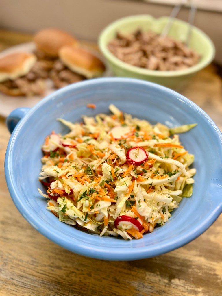 A big blue bowl of cabbage slaw with some pulled pork and sandwiches behind on a wood table
