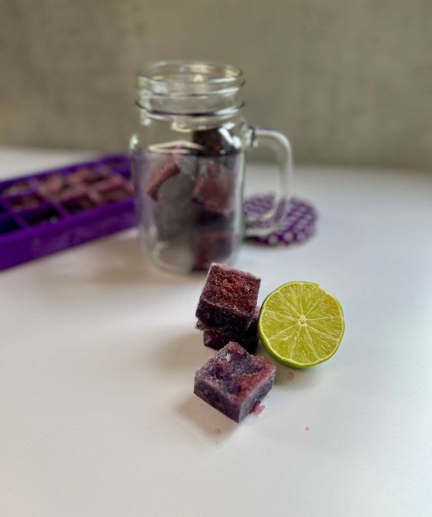 Boozy ice cubes stacked up with some lime next to it and a glass full of the cubes and an ice cube tray behind
