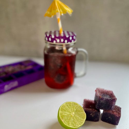 Boozy blackberry cubes stacked up with some lime next to it and a drink and ice cube tray behind
