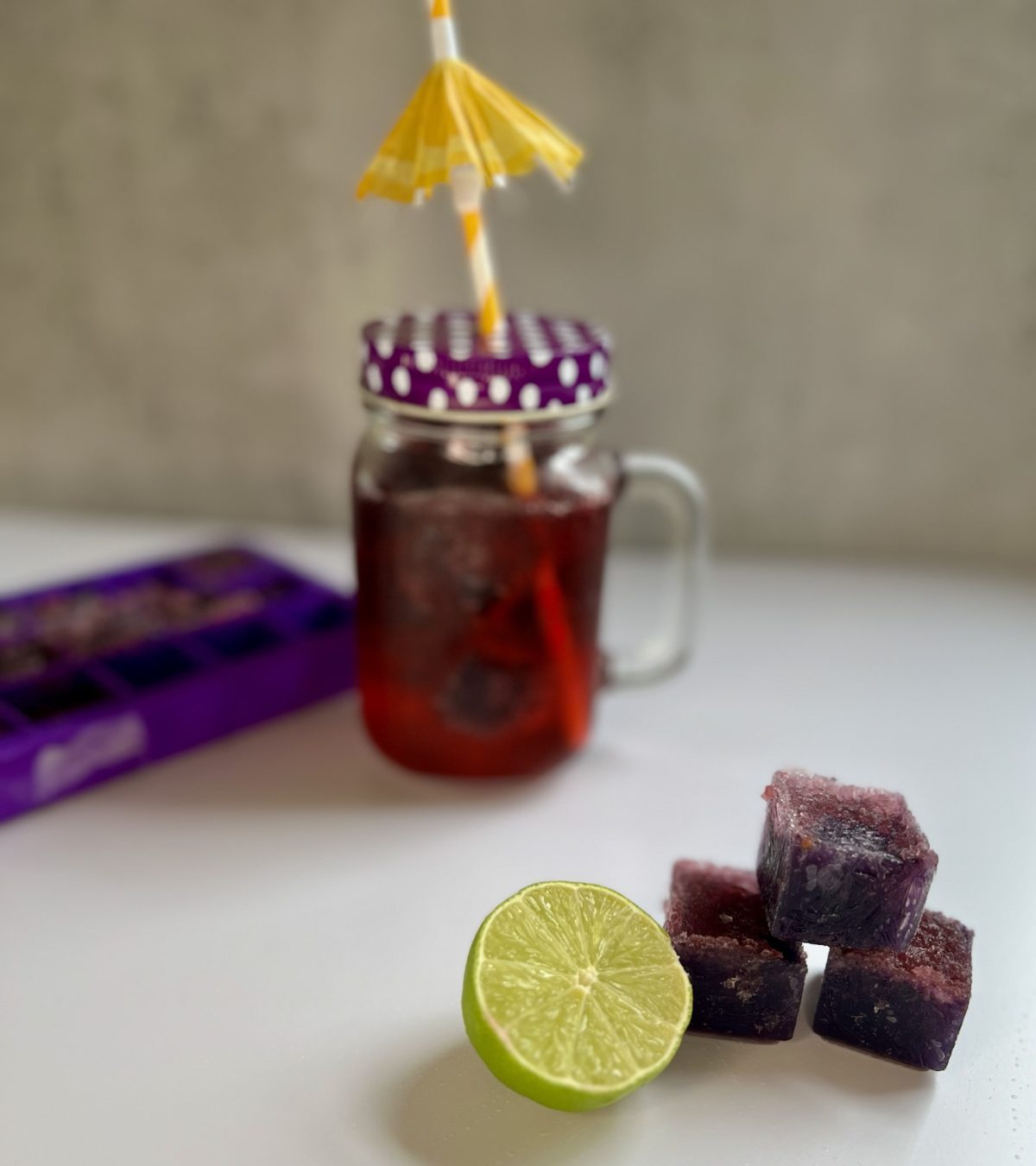 Boozy blackberry cubes stacked up with some lime next to it and a drink and ice cube tray behind