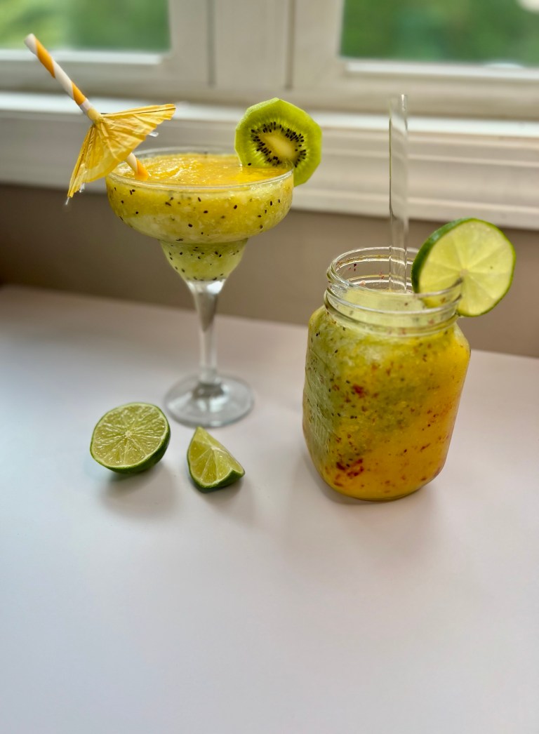 Kiwi Mango Daiquiri in a glass with a mason jar of it next to it with some kiwi and lime garnishes