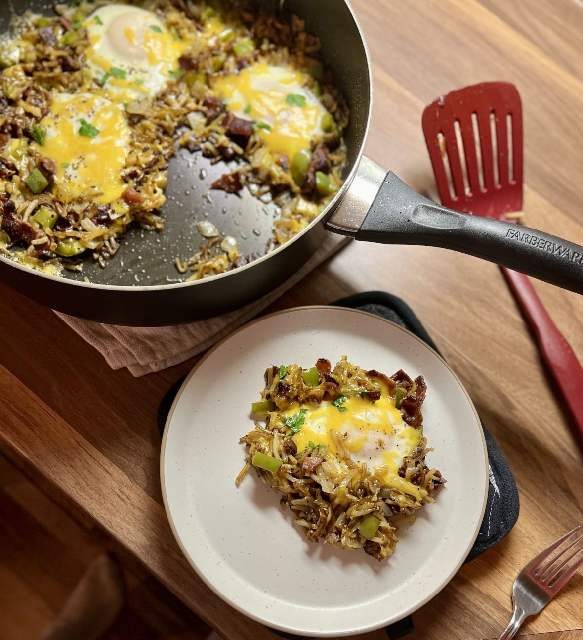 Bacon Breakfast Skillet in a skillet on a wooden counter with a white plate of it nearby and a spatula