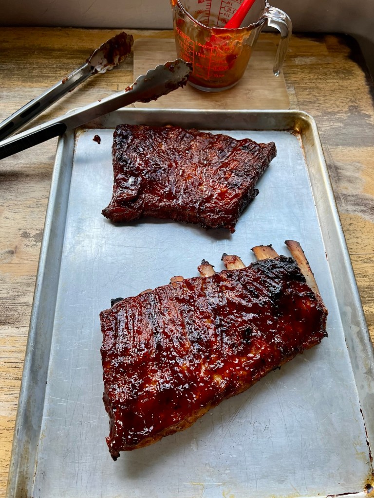 Grilled BBQ Ribs on a baking sheet with some sauce and tongs nearby