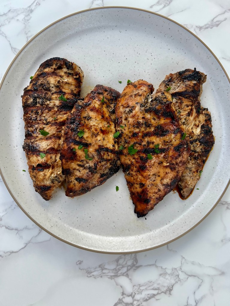 four chicken breasts that have been marinated and grilled on a white plate, on a marble countertop