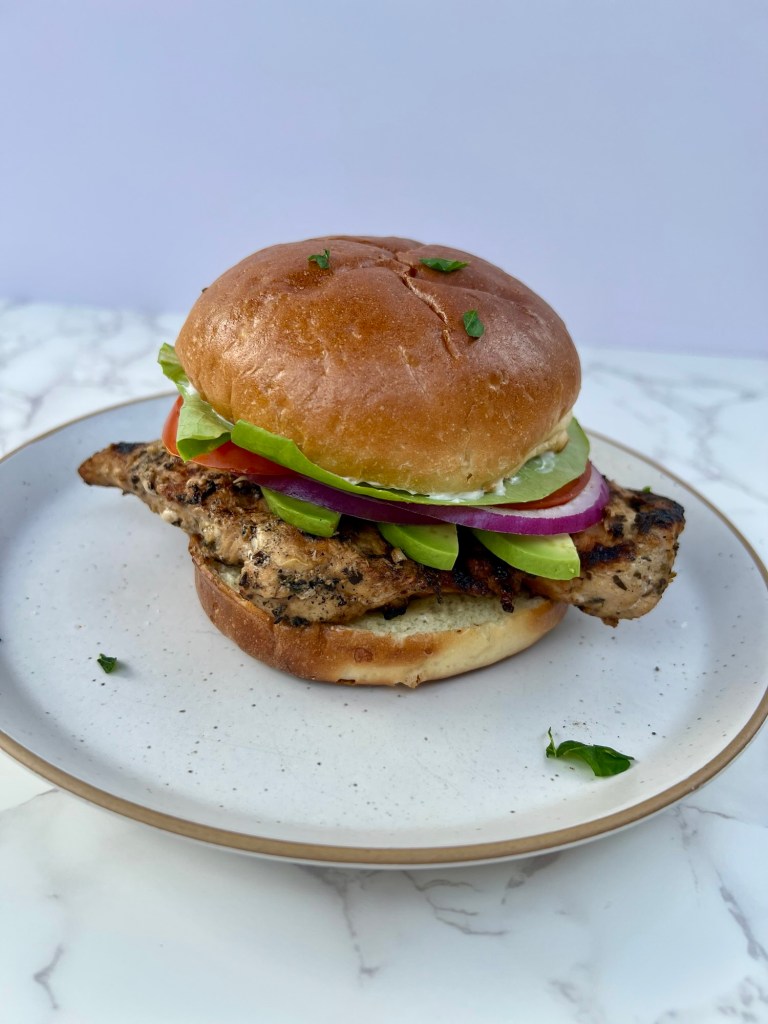 A grilled chicken sandwich on a white plate on a marble countertop