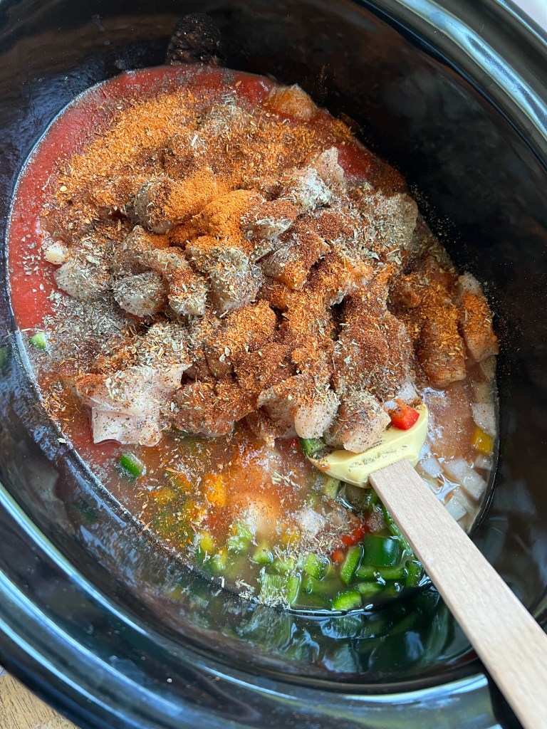 A crockpot filled with raw veggies, broth, seasoning, chicken, and sausage